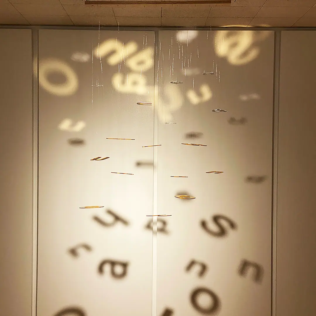 Suspended letters cast soft, scattered shadows onto a pale gallery wall