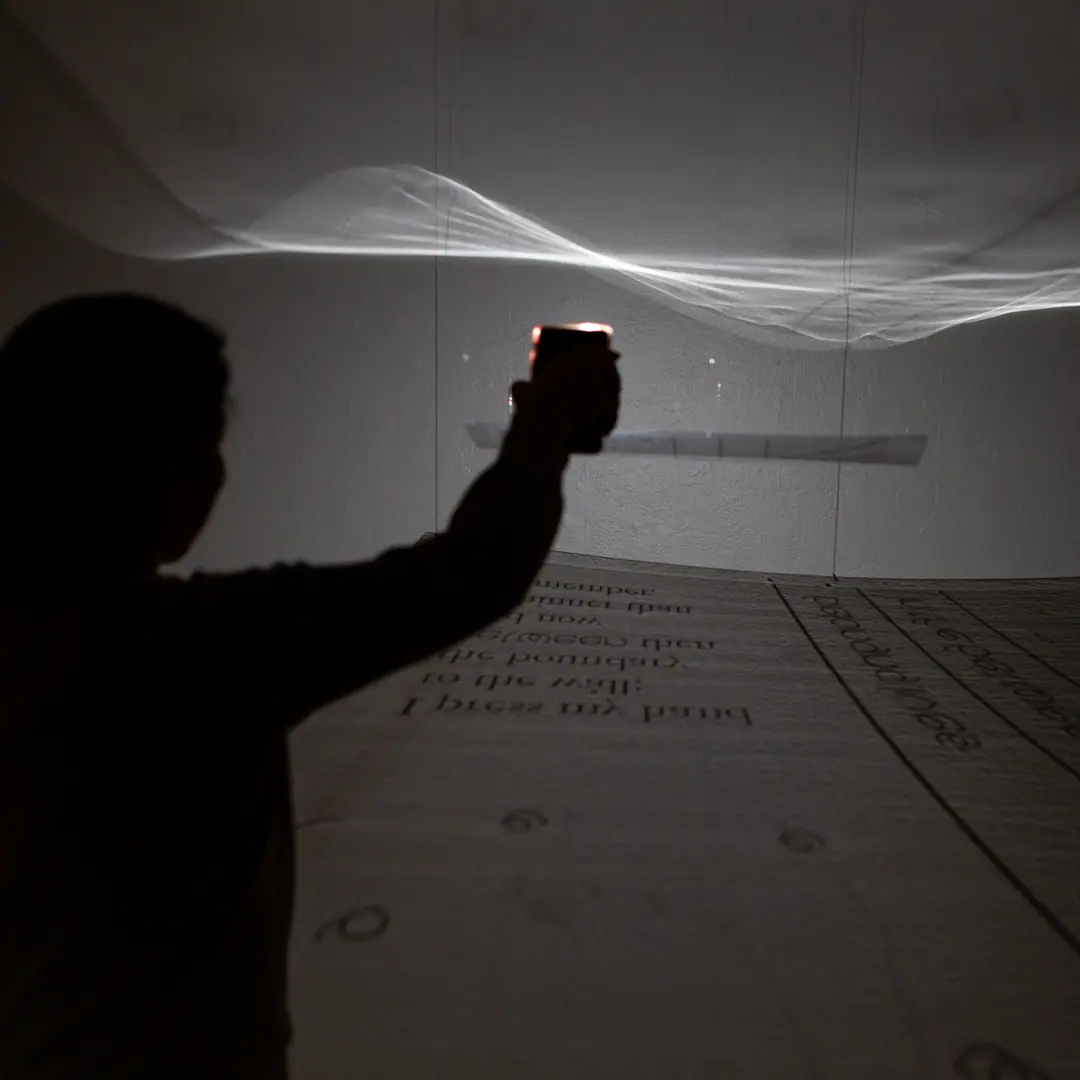 A visitor raises a light toward a suspended, translucent sheet of text that ripples like a wave in the dark