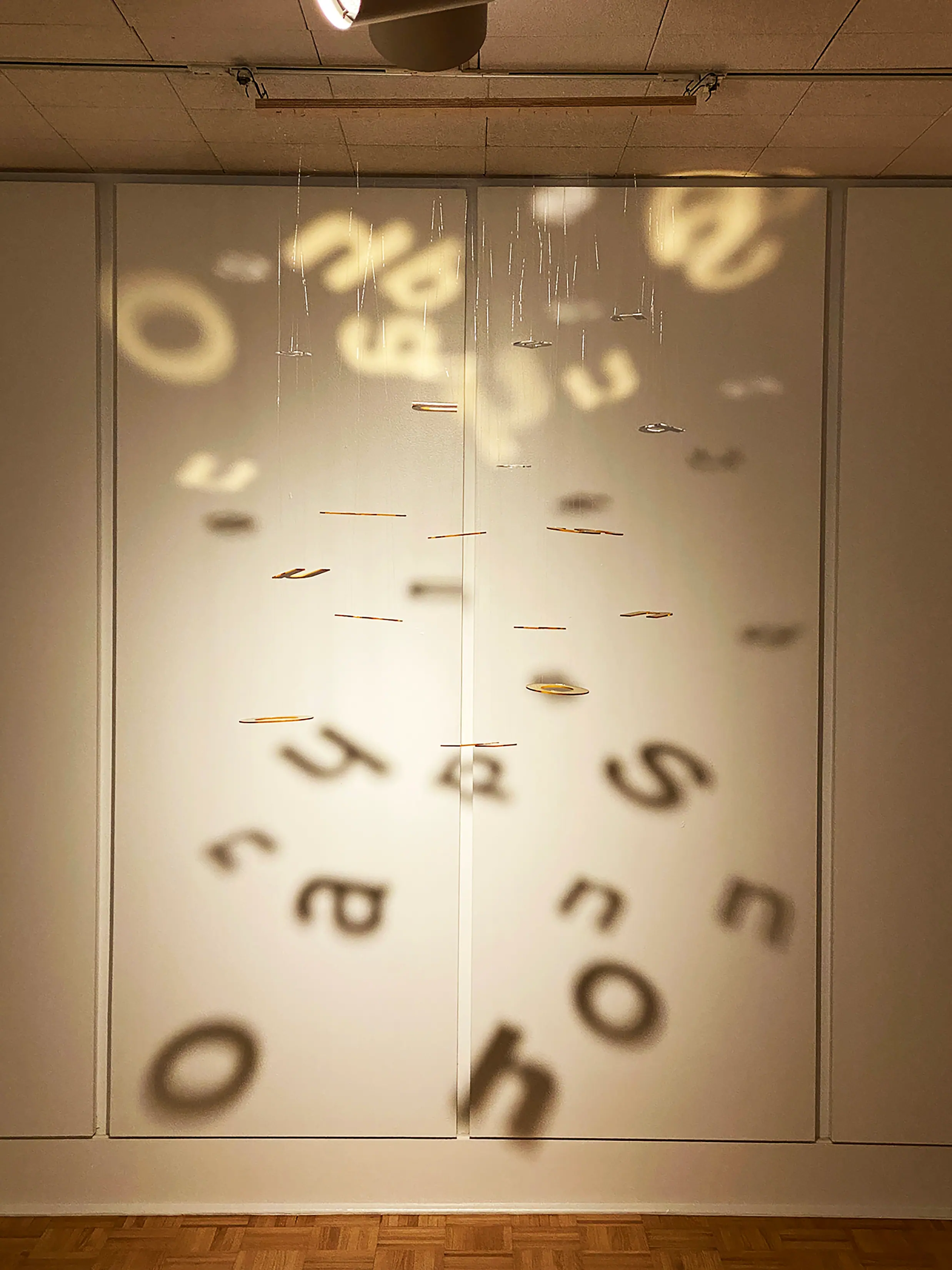 Suspended letters cast soft, scattered shadows onto a pale gallery wall