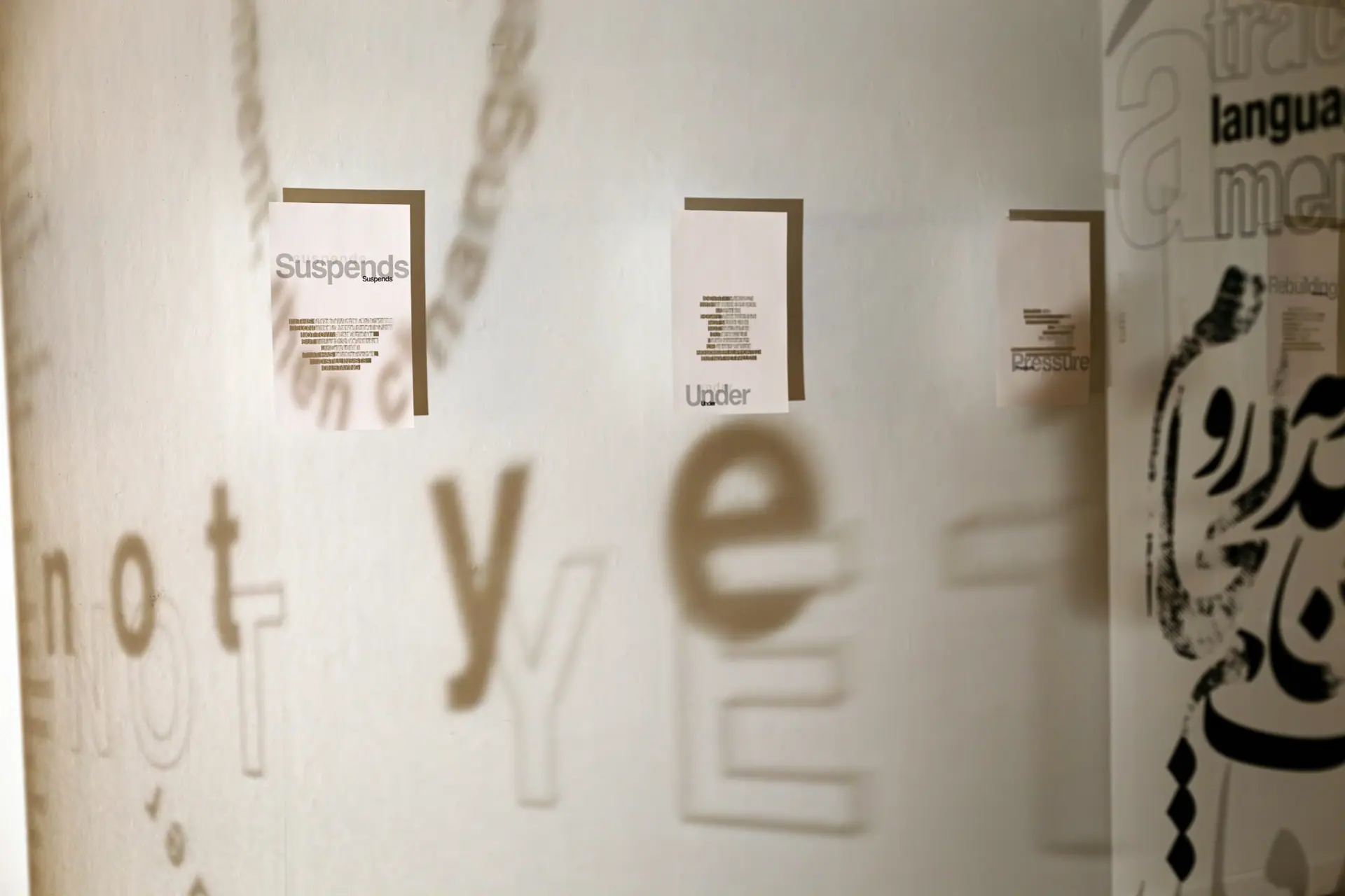 Shadows of the words 'not yet' and Persian calligraphy cast across a gallery wall between hanging text panels