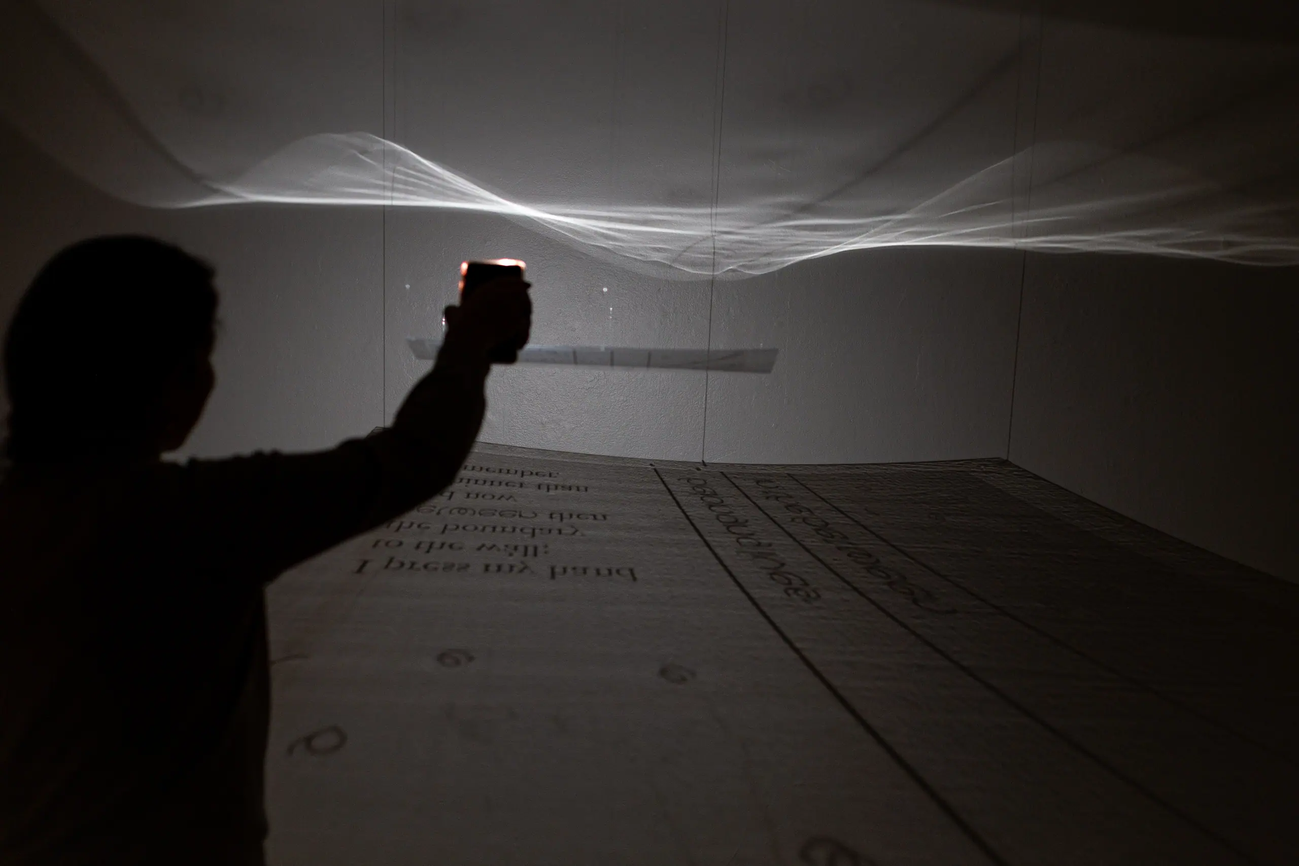 A visitor raises a light toward a suspended, translucent sheet of text that ripples like a wave in the dark