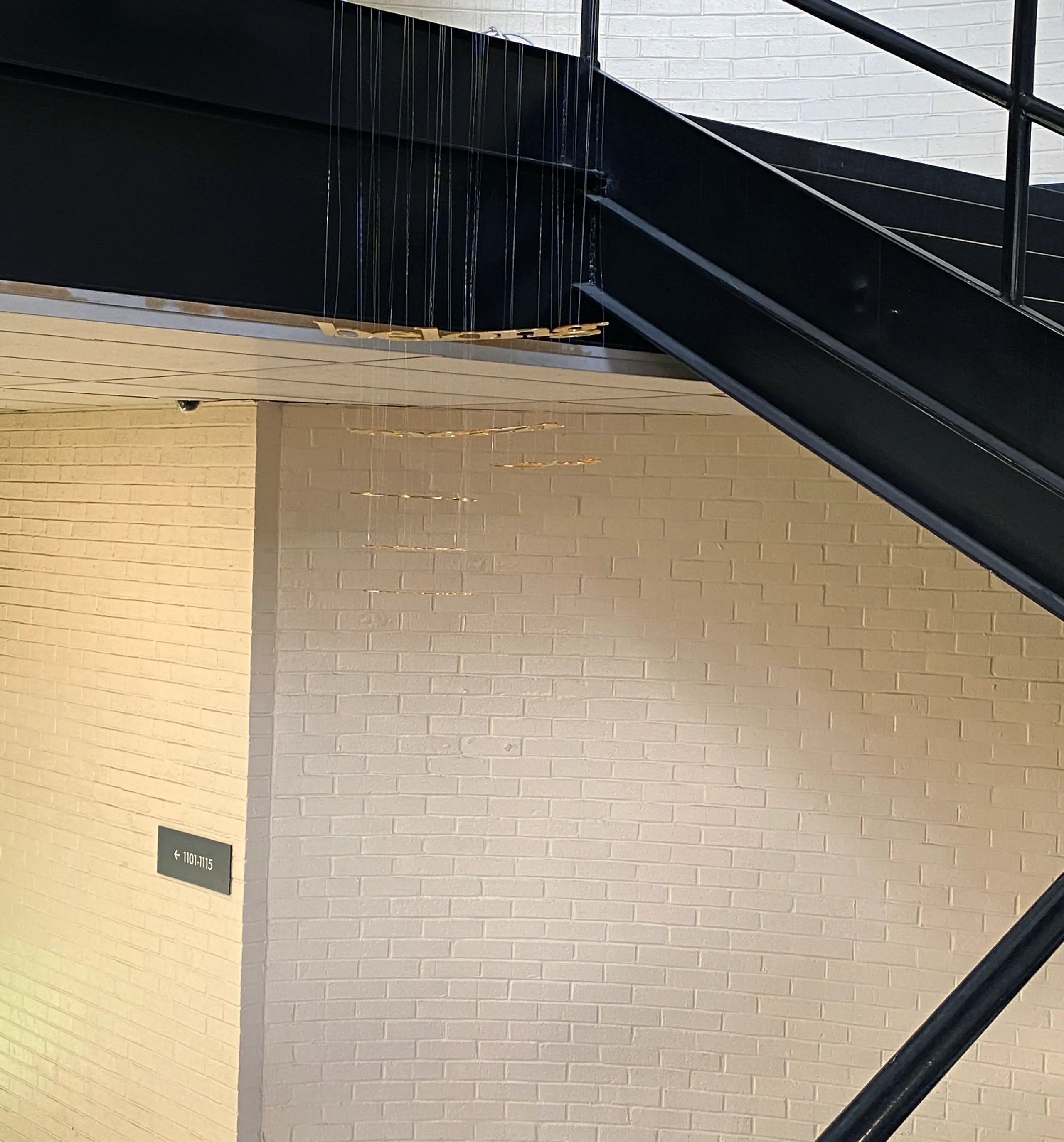 Gold cut letters hang on fine threads beneath a black steel staircase, in front of a white brick wall